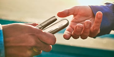 Hands exchanging IQOS Originals Duo heated tobacco holder and pocket charger in Brilliant Gold color.