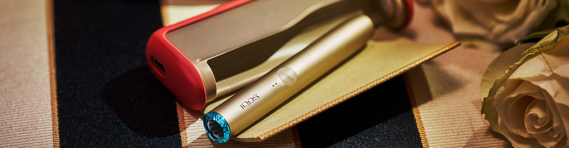 Podgrzewacz tytoniu IQOS ILUMA PRIME w złotym odcieniu z niebieską żłobioną nasadką leżący w otwartej przenośnej ładwarce w czerwonym etui]