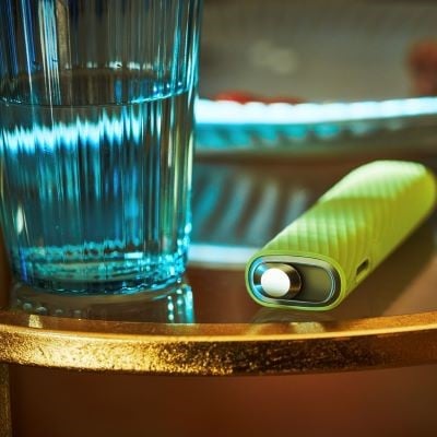 Podgrzewacz IQOS ILUMA ONE w silikonowej obudowie leżący obok szklanki z wodą