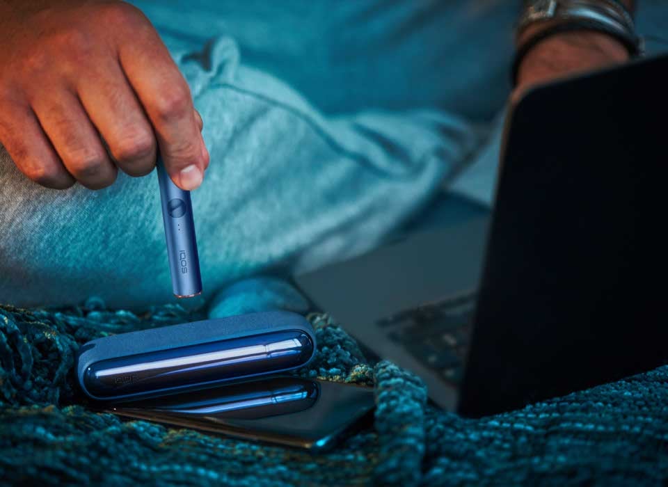 IQOS ILUMA w niebieskim odcieniu w dłoni osoby korzystającej z laptopa