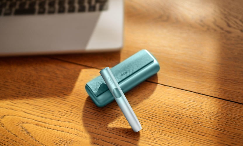 Podgrzewacz IQOS ILUMA i PRIME w kolorze Breeze Blue leżący na stole obok laptopa