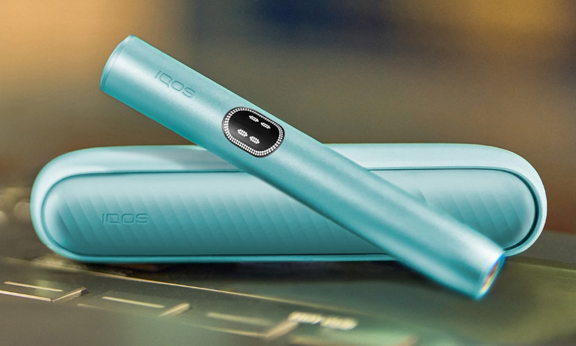 Podgrzewacz IQOS ILUMA i w błękitnym odcieniu oparty o leżącą obok przenośną ładowarkę
