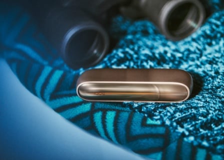 Brilliant gold IQOS 3 DUO charger on blue fabric next to binoculars