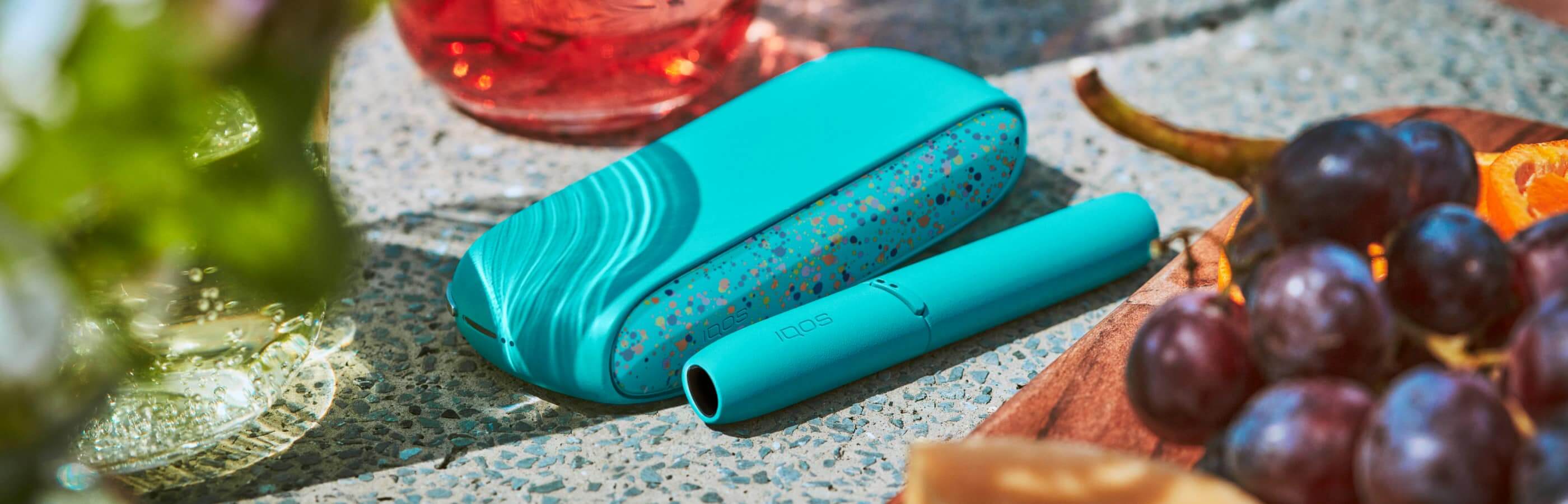 Ładowarka IQOS 3 DUO WE Edition z limitowanej serii w kolorze turkusowym z kolorowymi plamkami na nakładce obok podgrzewacza tytoniu przy napoju i misce owoców.