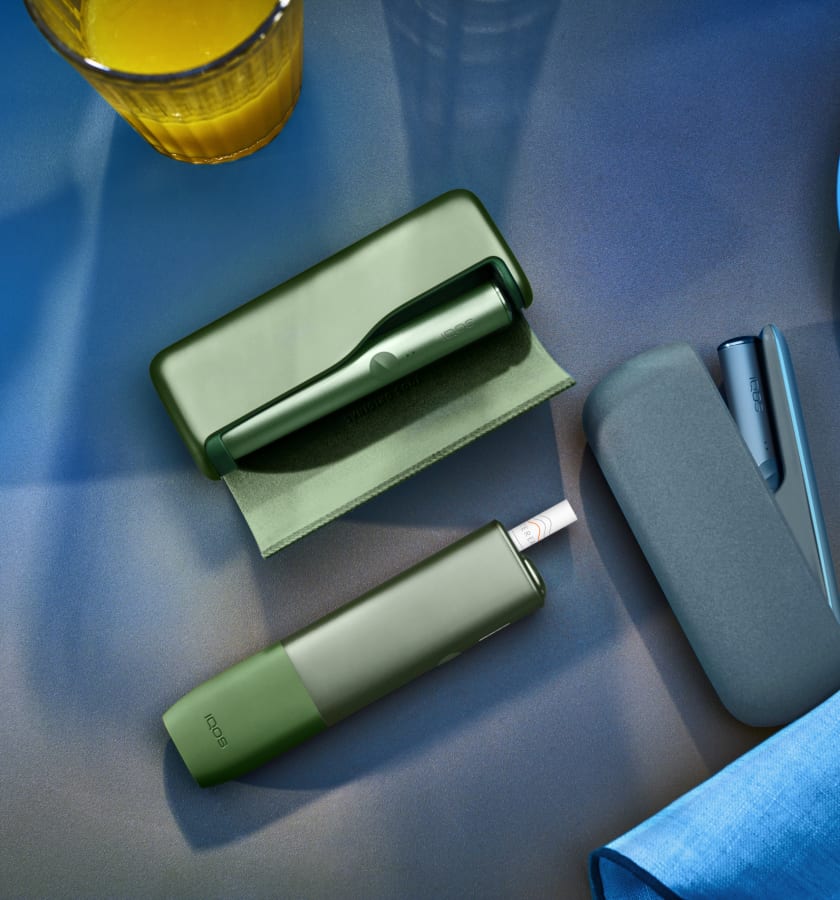 Podgrzewacz IQOS ILUMA leżący na stole obok innych modeli podgrzewaczy IQOS w kolorze Moss Green