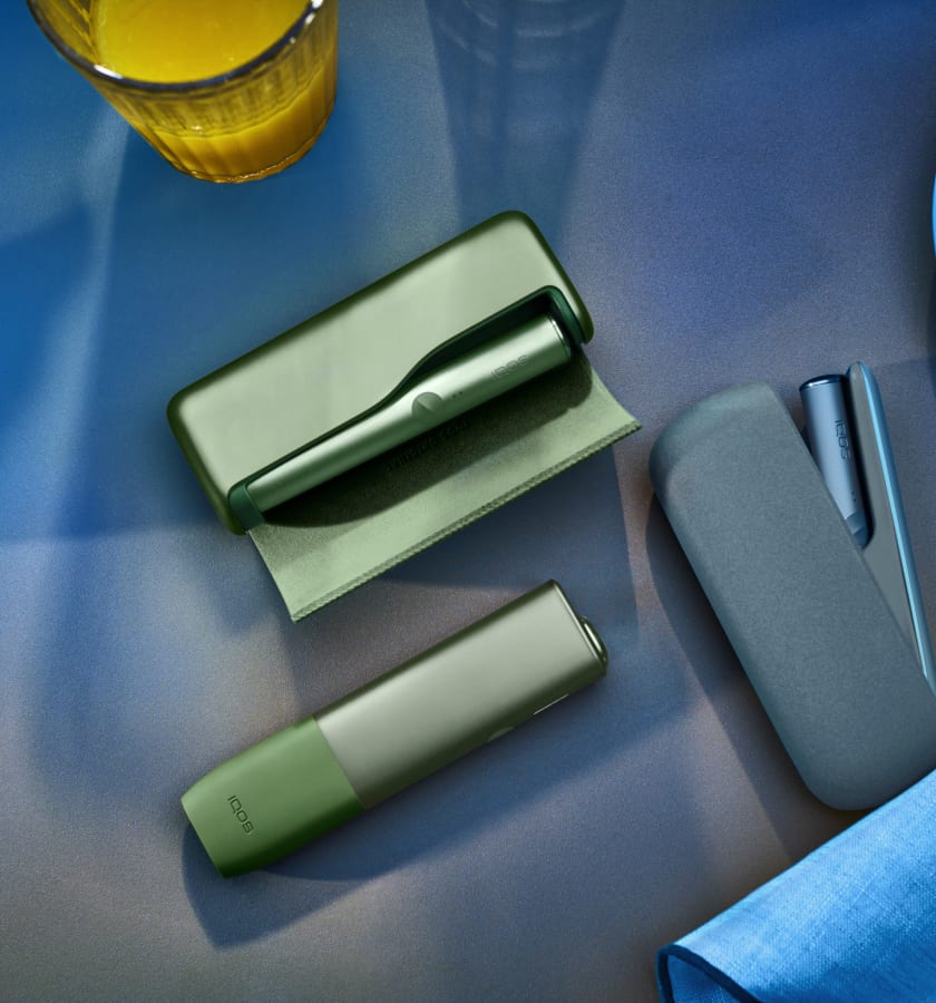 Podgrzewacz IQOS ILUMA leżący na stole obok innych modeli podgrzewaczy IQOS w kolorze Moss Green