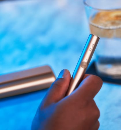 Podgrzewacz IQOS ILUMA PRIME trzymany w dłoni w tle przenośna ładowarka leżąca obok szklanki z wodą