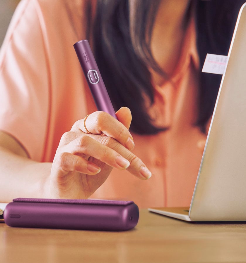 IQOS ILUMA PRIME w kolorze Obsidian Black schowany w przenośnej ładowarce trzymany w dłoni przez osobę korzystającą z laptopa
