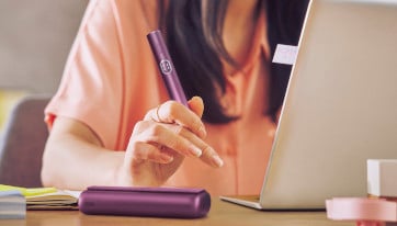 Podgrzewacz IQOS ILUMA i PRIME w dłoni osoby siedzącej przy biurki przy laptopie