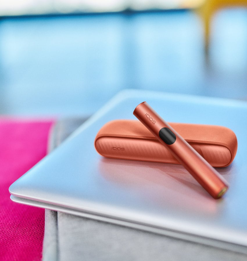 Podgrzewacz IQOS ILUMA i z przenośną ładowarką w czerwony odcieniu leżące na zamkniętym laptopie
