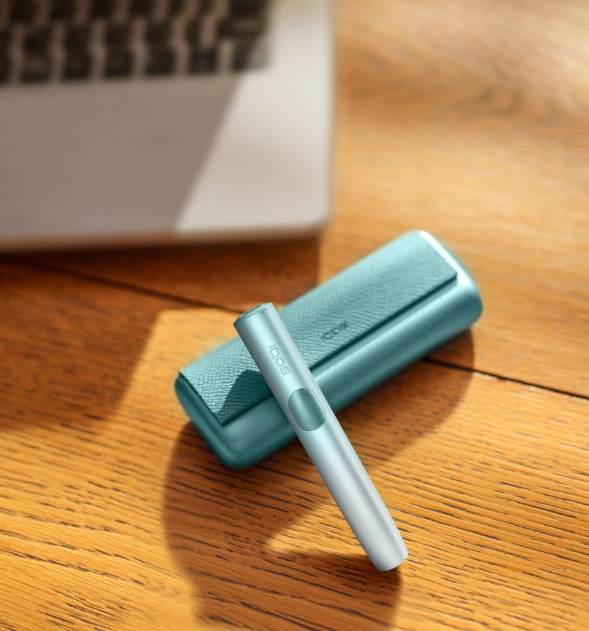Podgrzewacz IQOS ILUMA i PRIME w kolorze Breeze Blue leżący na stole obok laptopa