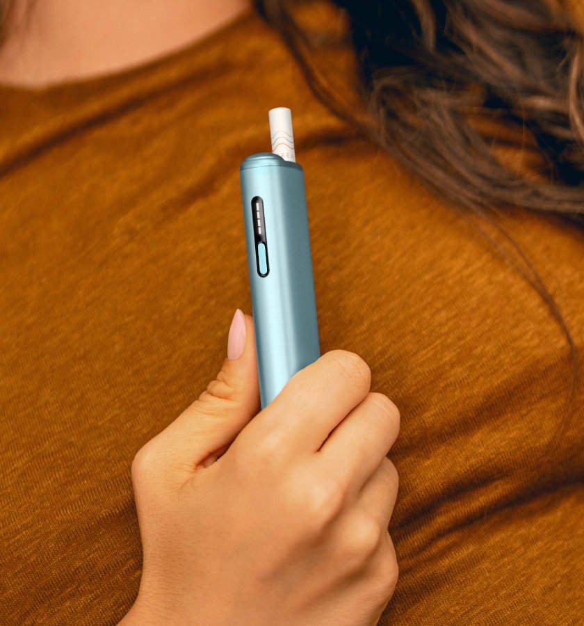Podgrzewacz tytoniu IQOS ILUMA i ONE w kolorze Breeze Blue, z wsuniętym wkładem tytoniowym, trzymany w dłoni