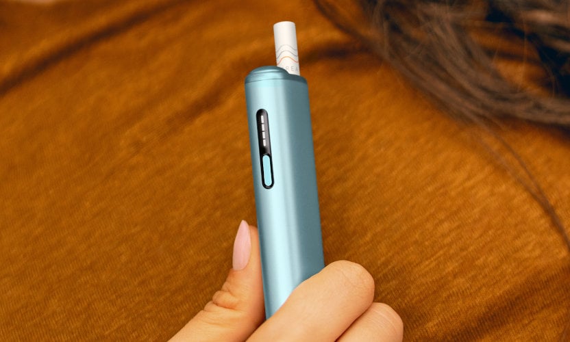 Podgrzewacz tytoniu IQOS ILUMA i ONE w kolorze Breeze Blue, z wsuniętym wkładem tytoniowym, trzymany w dłoni