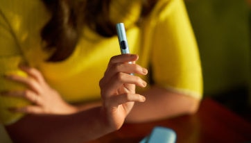 Podgrzewacz IQOS ILUMA i trzymany w dłoni przez kobietę w żółtej bluzce