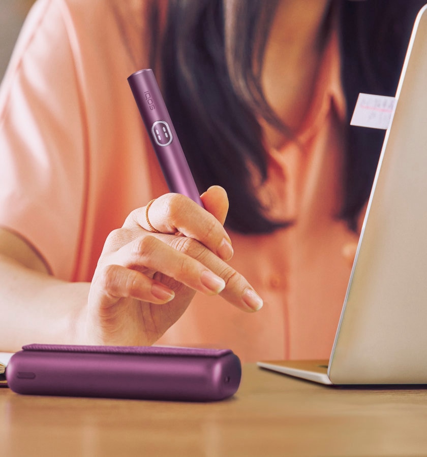 IQOS ILUMA PRIME w kolorze Obsidian Black schowany w przenośnej ładowarce trzymany w dłoni przez osobę korzystającą z laptopa