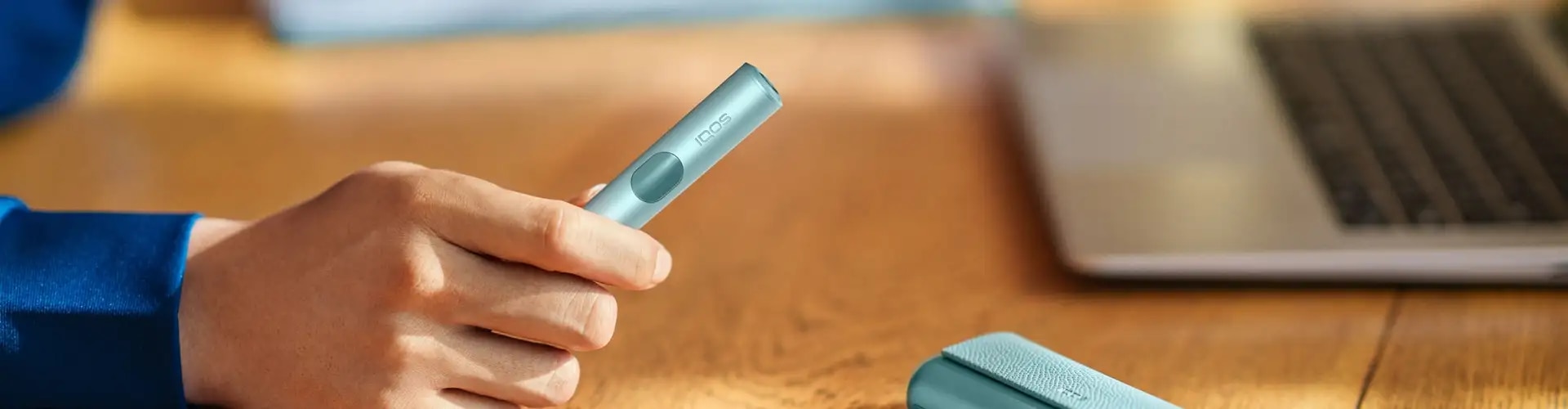Podgrzewacz IQOS ILUMA i PRIME, błękitny, trzymany w dłoni, w tle laptop 