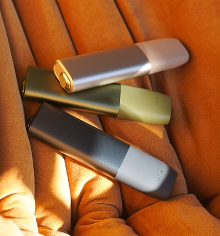 3 IQOS-y ILUMA ONE w różnych kolorach leżą na pofałdowanej, miękkiej tkaninie