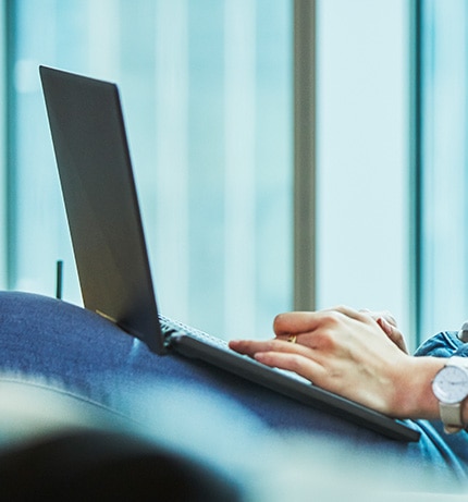 Otwarty laptop widoczny bokiem na kolanach osoby, której dłonie widzimy na jego klawiaturze