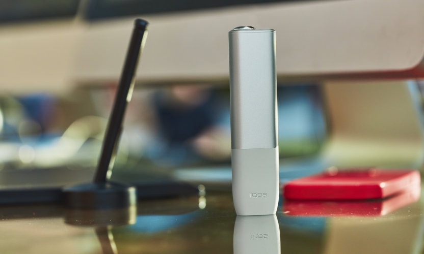 Beżowy IQOS ILUMA ONE stoi na biurku w otoczeniu elektornicznych urządzeń biurowych