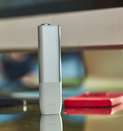 Beżowy IQOS ILUMA ONE stoi na biurku w otoczeniu elektornicznych urządzeń biurowych