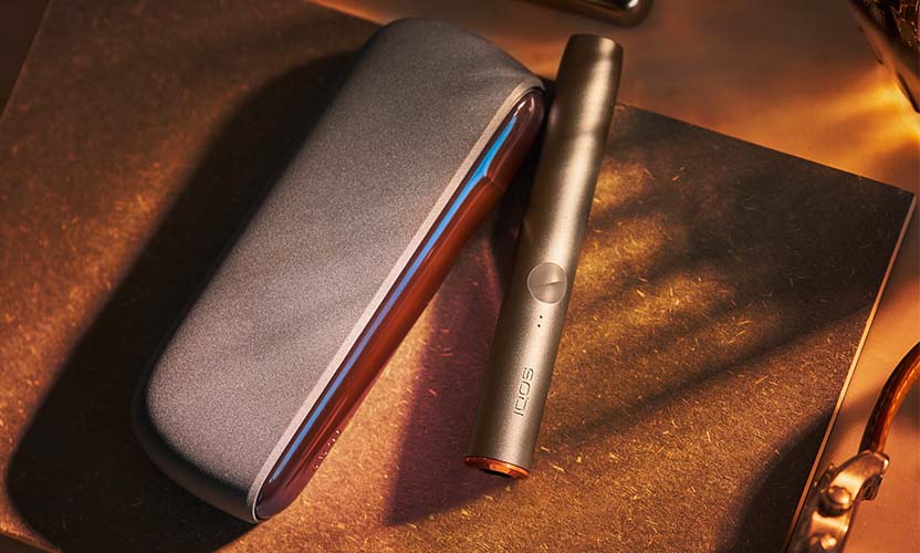 Ładowarka i podgrzewacz IQOS ILUMA w kolorze beżowym, z wymienioną nakładką na ładowarce i nasadą na podgrzewaczu, leżą na skórzanej podkładce