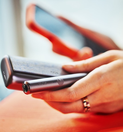 Podgrzewacz i ładowarka IQOS ILUMA PRIME w kolorze beżowym w jednej dłoni. W tle telefon w drugiej dłoni