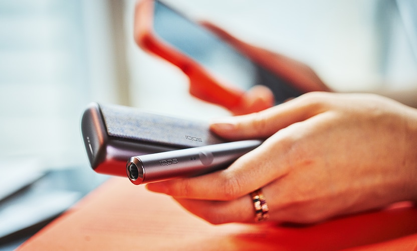 Podgrzewacz i ładowarka IQOS ILUMA PRIME w kolorze beżowym w jednej dłoni. W tle telefon w drugiej dłoni