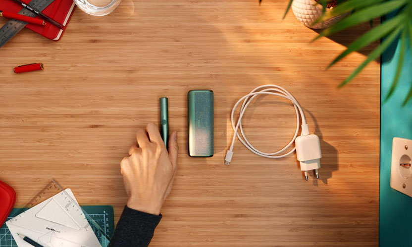 Widok z góry na biurko, na którym leży podgrzewacz i ładowarka IQOS ILUMA PRIME w kolorze zielonym i kabel do ładowania. Dłoń sięga po podgrzewacz.