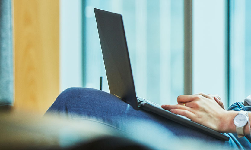 Otwarty laptop widoczny bokiem na kolanach osoby, której dłonie widzimy na jego klawiaturze