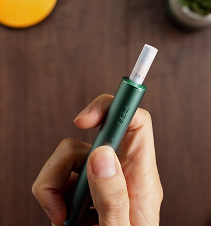 IQOS ILUMA PRIME w kolorze zielonym z wkładem. Podgrzewacz w dłoni z kciukiem na przycisku. Widok z góry na tle drewnianego blatu.