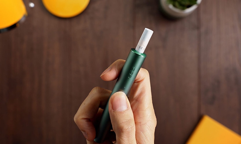 IQOS ILUMA PRIME w kolorze zielonym z wkładem. Podgrzewacz w dłoni z kciukiem na przycisku. Widok z góry na tle drewnianego blatu.