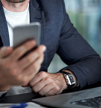 Mężczyzna w marynarce siedzi przy biurku z otwartym laptopem. W dłoni trzyma telefon