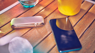 Gold IQOS DUO with a mobile, a glass of water and a yellow cup on a wooden table