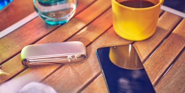 Gold IQOS device, a mobile, a yellow cup and a glass of water on a table bar 