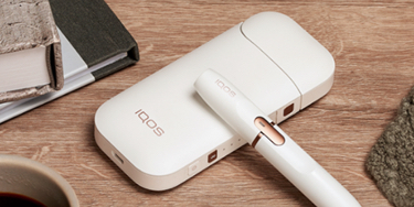 White IQOS 2.4 PLUS holder and charger on a wooden table next to a cup