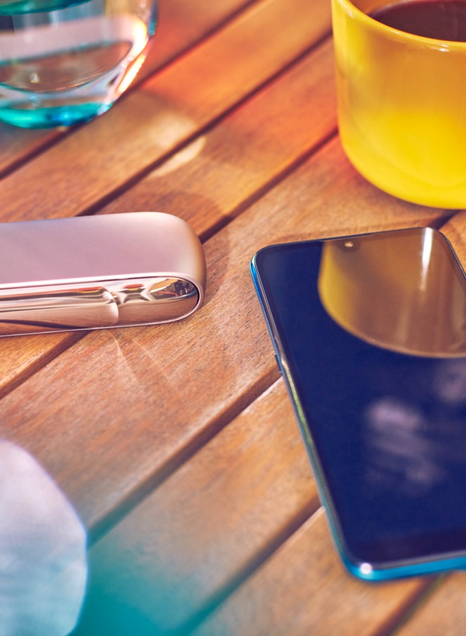 Gold IQOS device, a mobile, a yellow cup and a glass of water on a table bar 