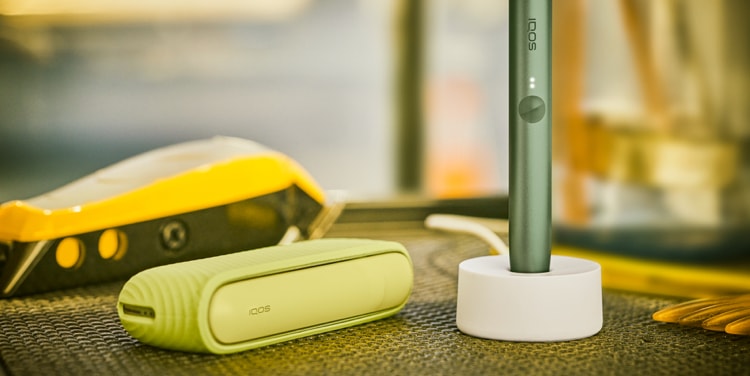 An IQOS ILUMA device in a charging station with a Pocket Charger on a table