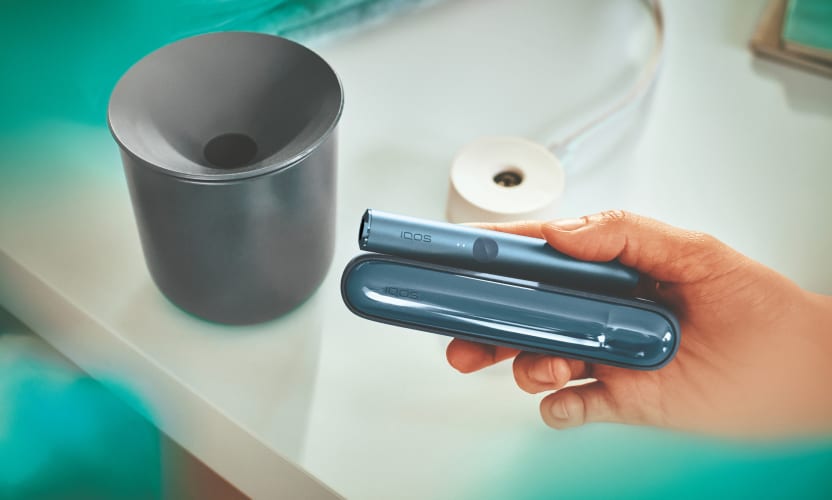Podgrzewacz IQOS ILUMA trzymany w dłoni w tle pojemnik na zużyte wklady
