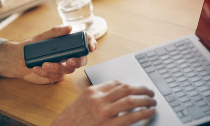 IQOS ILUMA PRIME w kolorze Obsidian Black schowany w przenośnej ładowarce trzymany w dłoni osoby korzystającej z laptopa