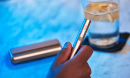 Podgrzewacz IQOS ILUMA PRIME trzymany w dłoni w tle przenośna ładowarka leżąca obok szklanki z wodą
