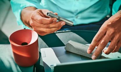 Trzymane w dłoniach podgrzewacz IQOS ILUMA PRIME I ładowarka, obok stojący pojemnik IQOS na zużyte wkłady