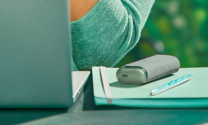Podgrzewacz IQOS ILUMA PRIME lezacy na notesie obok osoby korzystającej z laptopa